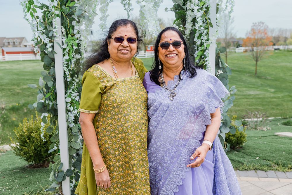 Two women in traditional Indian saris pose together in an outdoor garden setting with a floral arch.