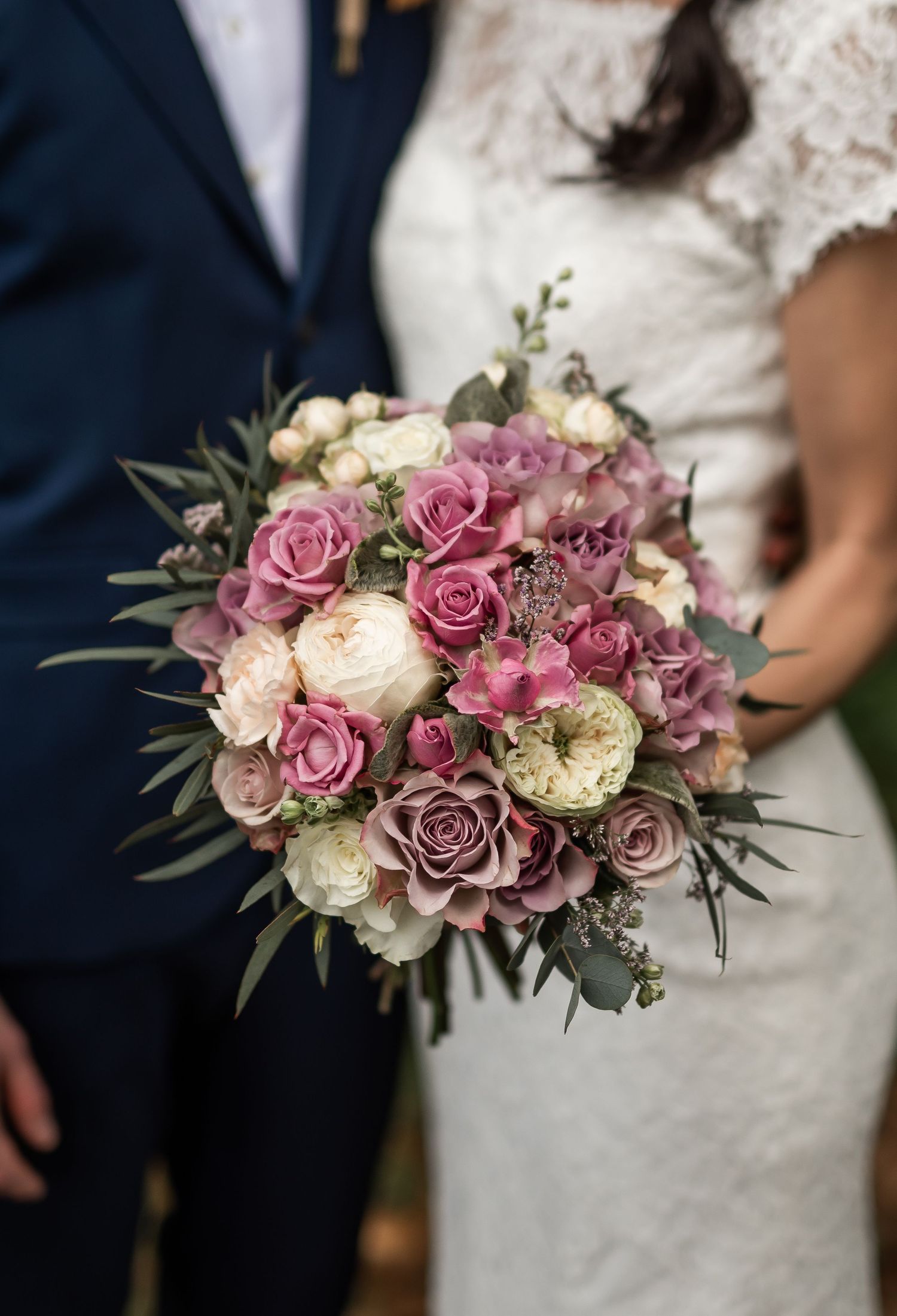 Elegant bridal bouquet of dusty pink, mauve, and cream roses held by a couple in wedding attire outdoors.