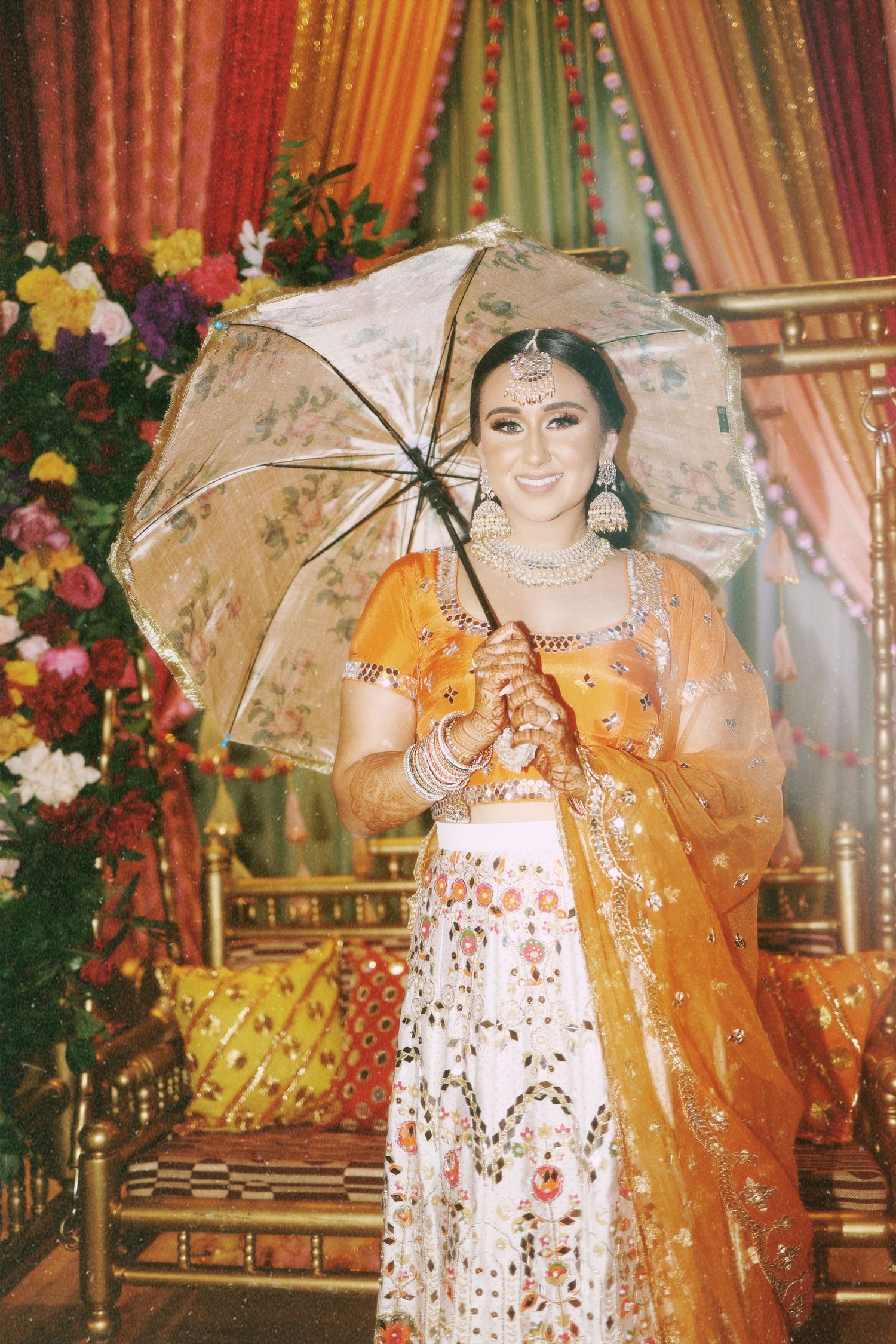 A bride in gold and white attire holds a decorative umbrella against colorful draped backdrop.