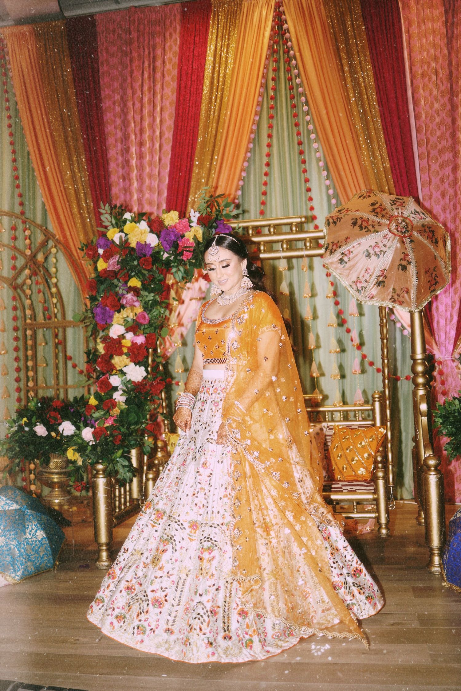 A bride in a golden lehenga poses in front of colorful draped fabric and floral decorations.