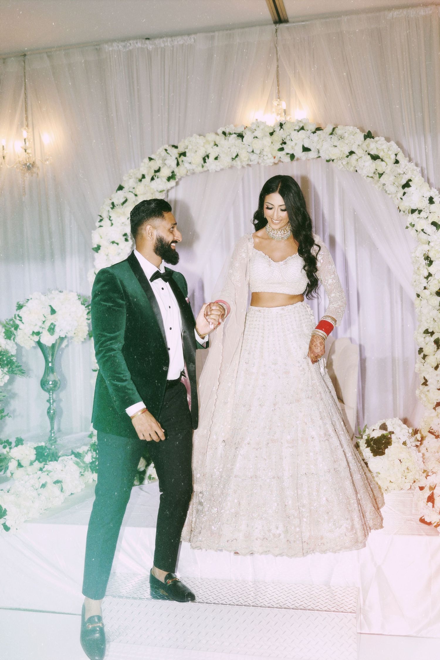 Wedding couple under floral arch, groom in emerald suit and bride in white two-piece lehenga.
