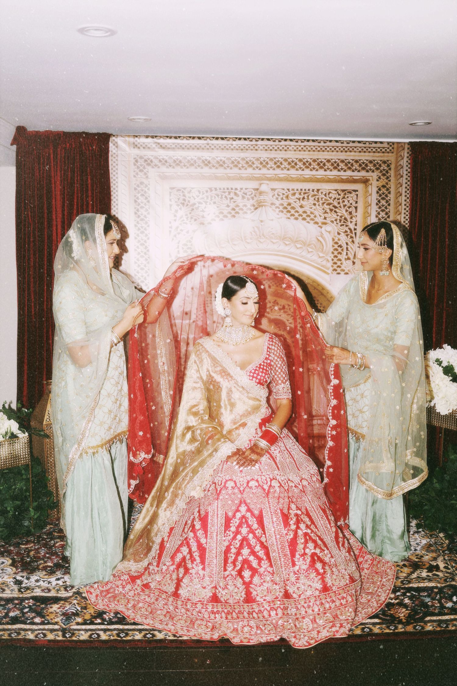 Traditional Indian bridal preparation scene with red lehenga and attendants in white.