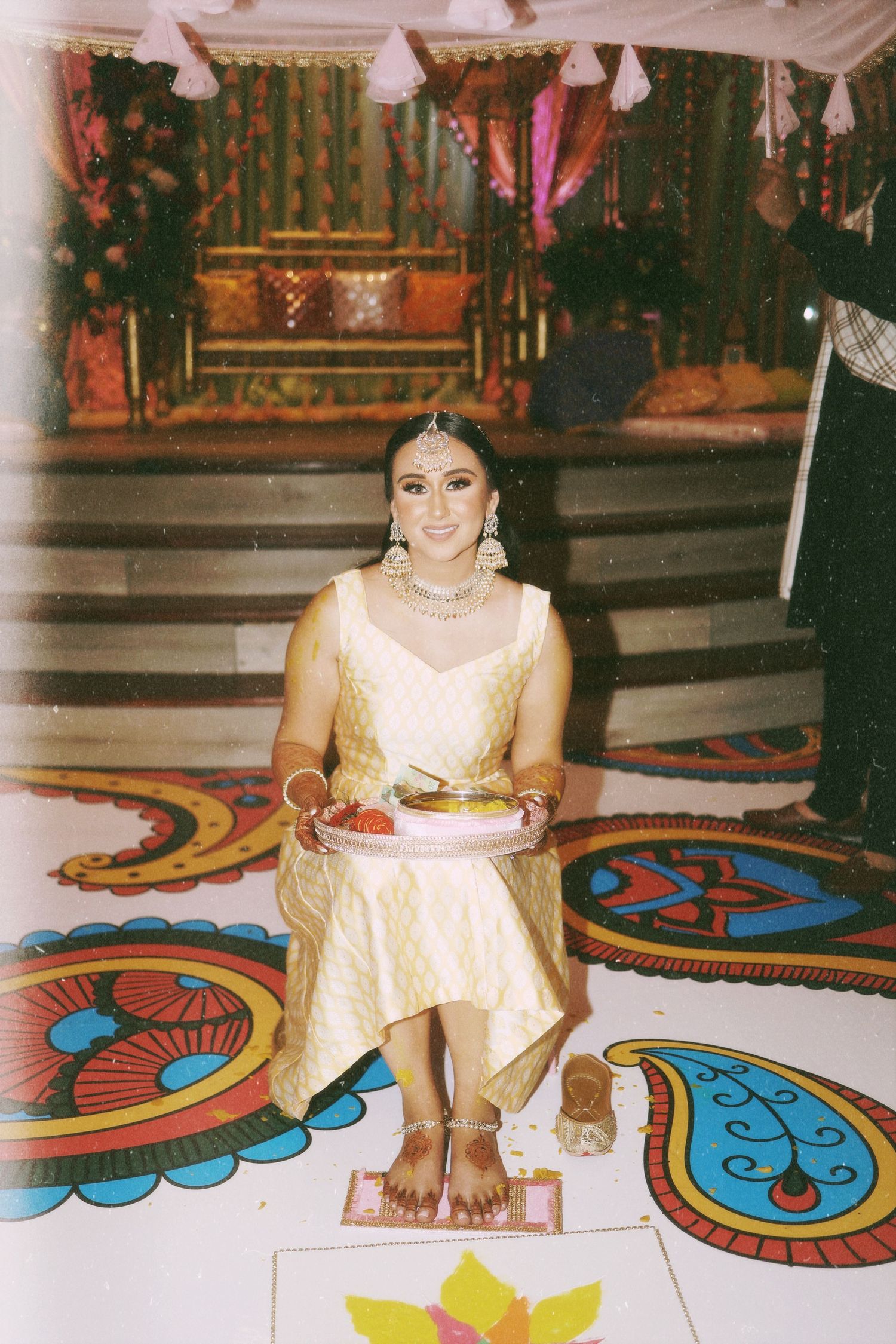 A person wearing a cream-colored traditional Indian outfit sits on a decorative floor with colorful paisley patterns.