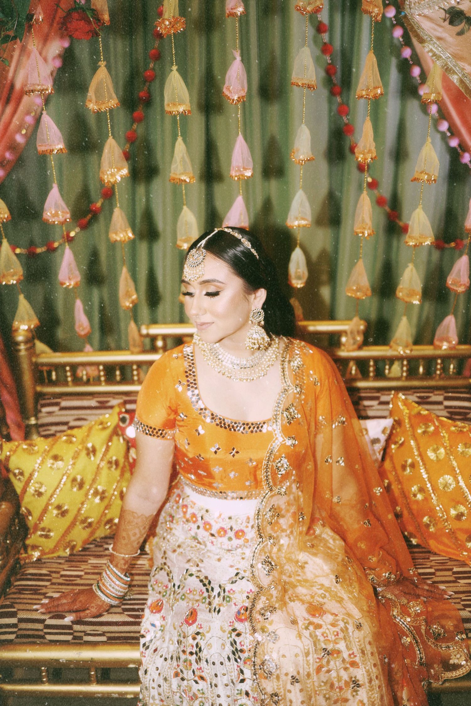 A bride wearing an orange and white lehenga poses against hanging decorative tassels.