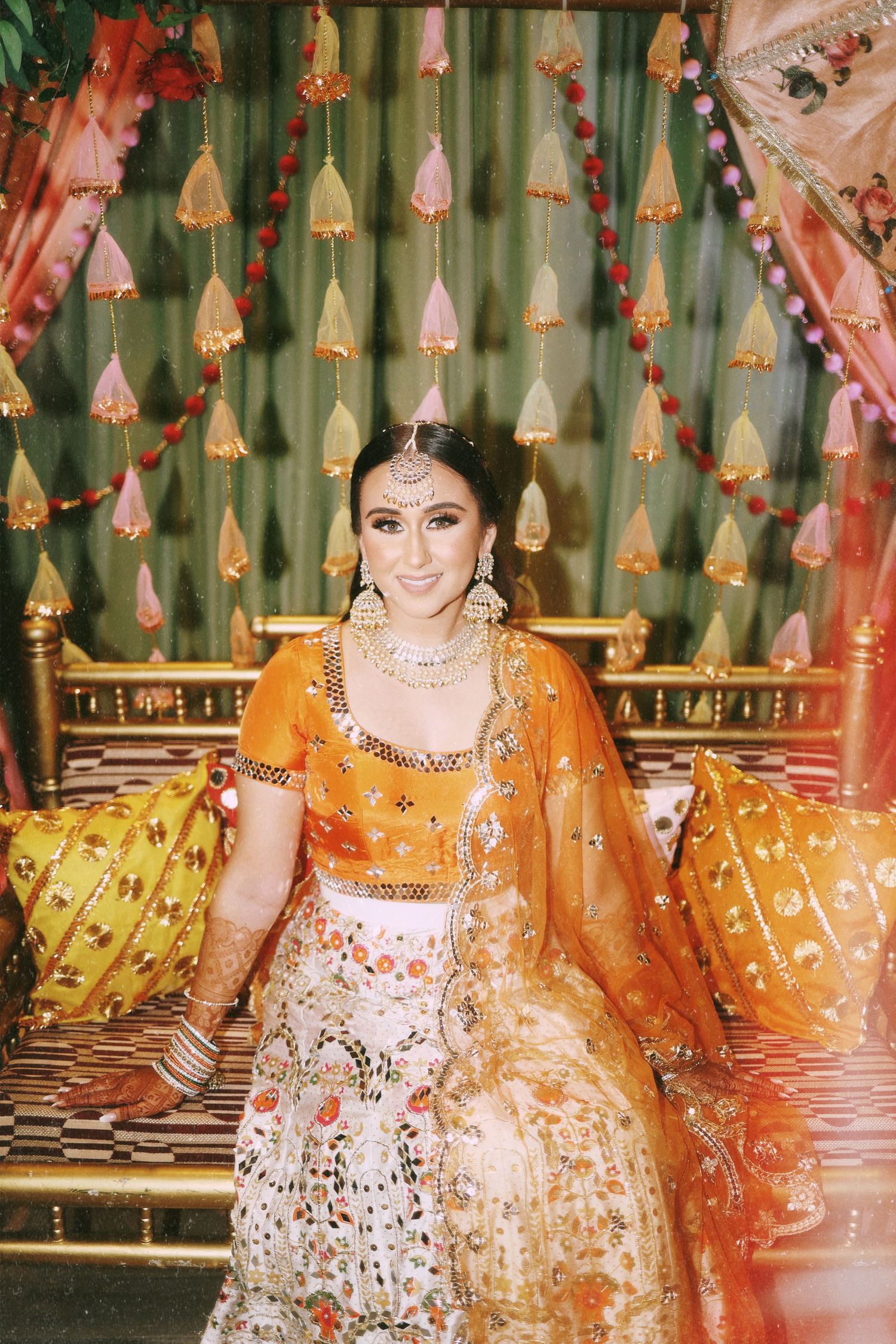 A bride in orange and white lehenga sits under hanging tassels against a green backdrop.