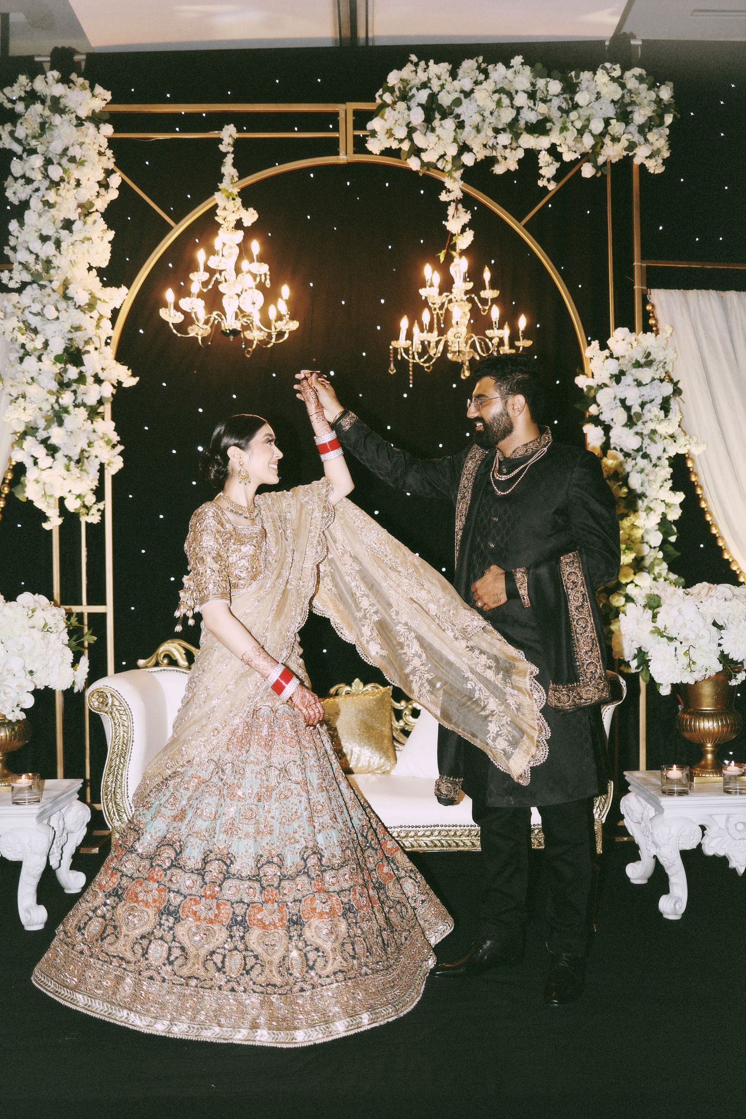 A couple in ornate wedding attire poses beneath a white floral arch with chandeliers.