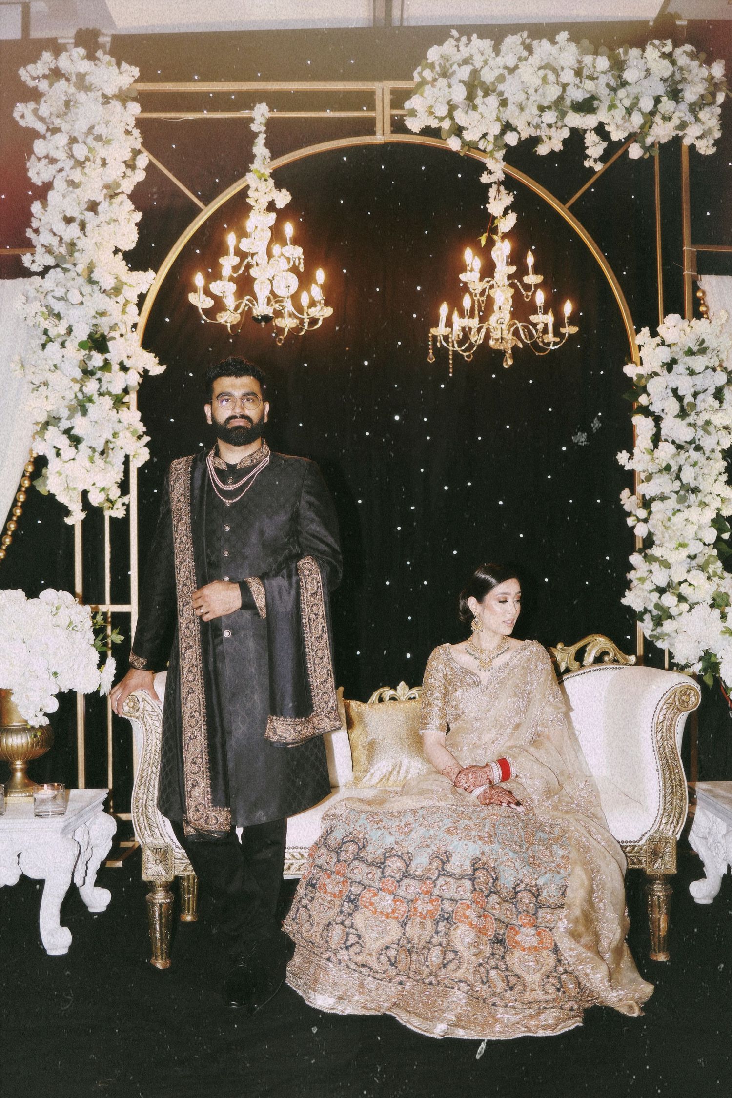 A couple poses under an elegant white floral arch with crystal chandeliers at their wedding.