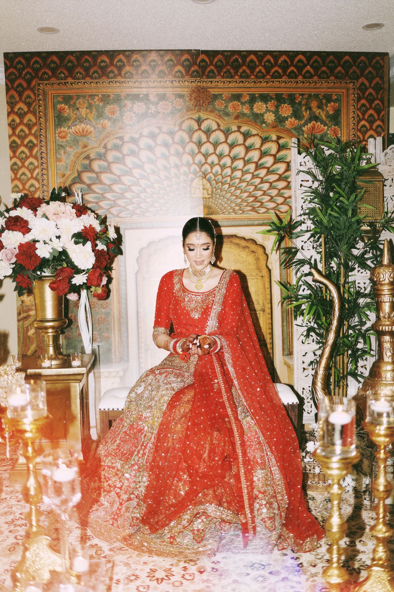 Traditional Indian bridal attire featuring an ornate red and gold lehenga against an intricately patterned backdrop.