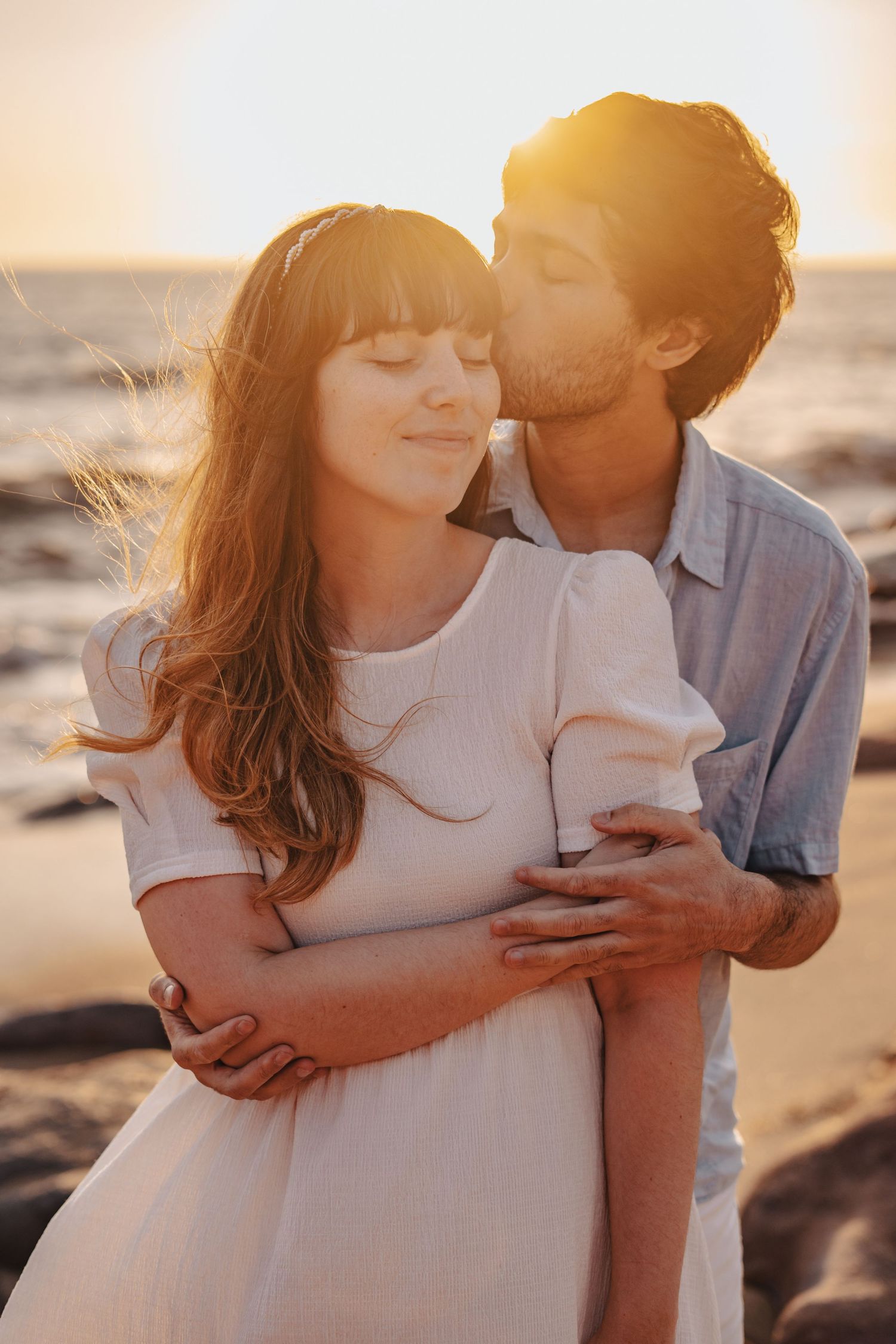 Romantic couple embraces on beach at sunset with soft golden light creating intimate atmosphere.