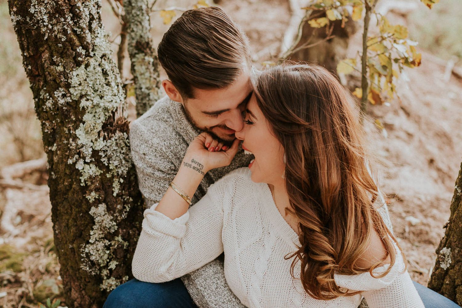 Couple sharing laughter while leaning against a tree trunk in warm sunlight.