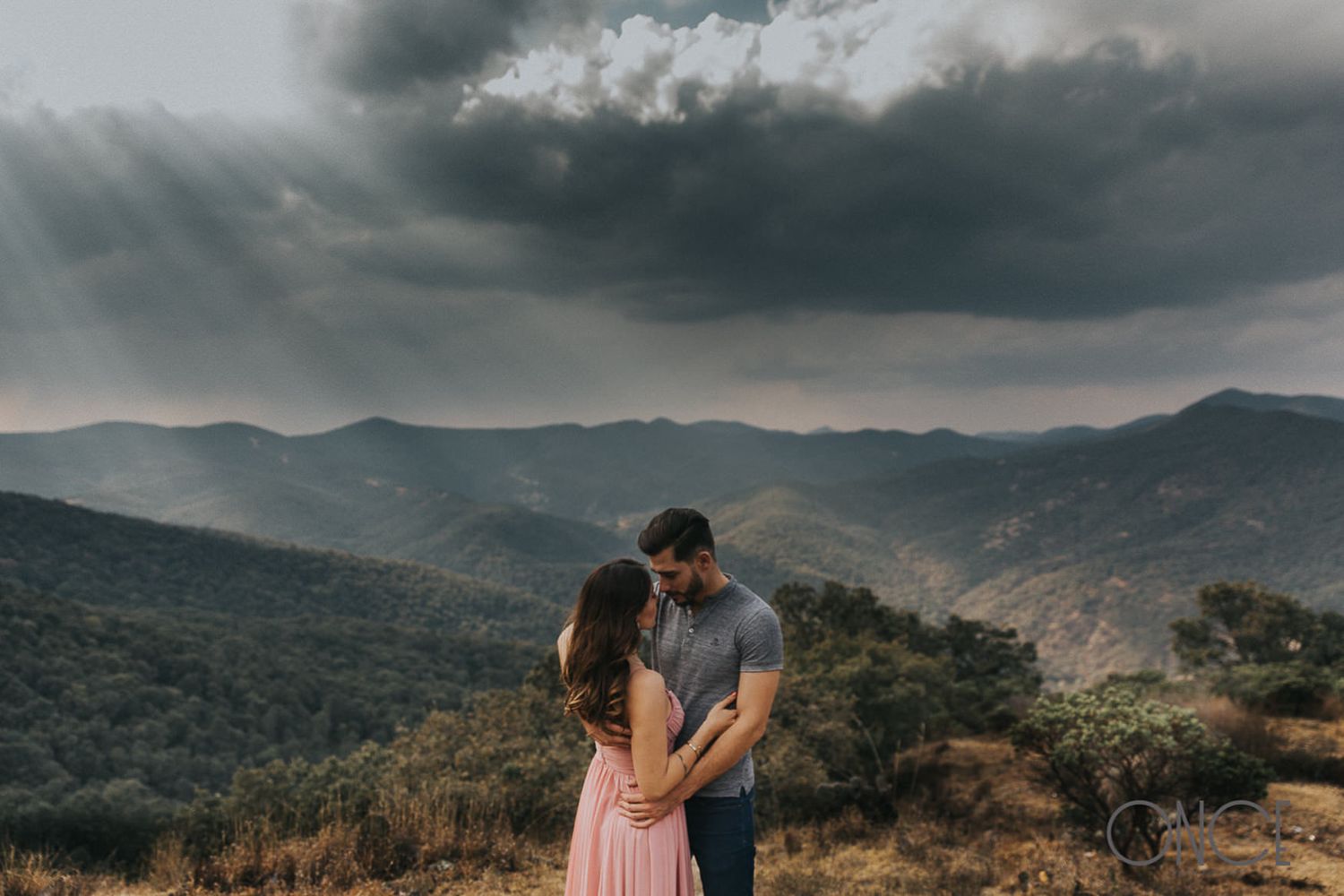 Couple embracing on mountaintop with dramatic stormy sky and valley view.