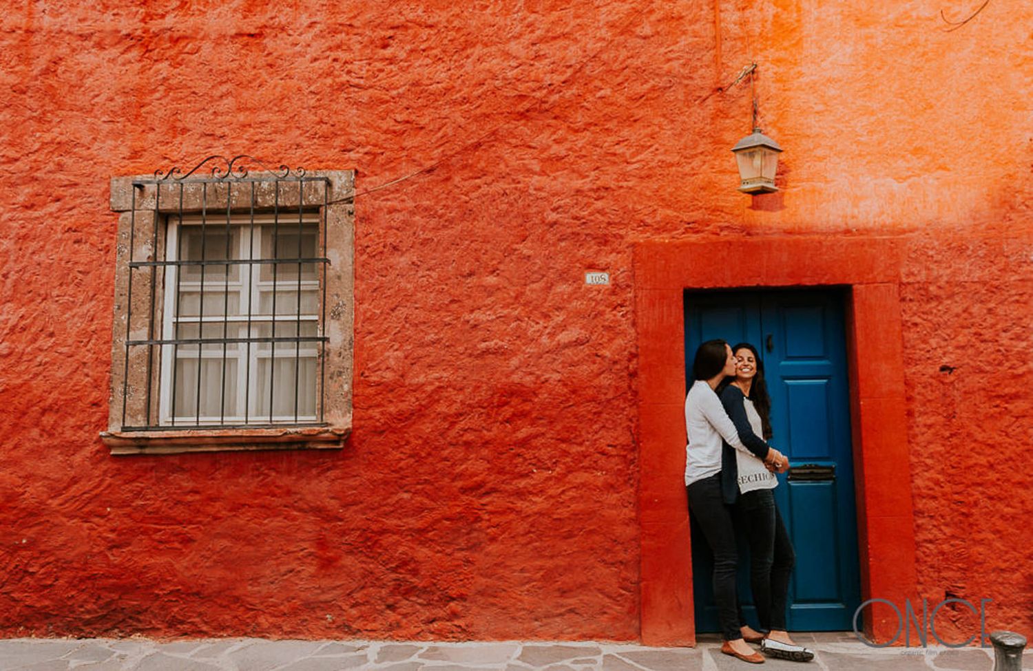 Person standing against bright orange wall with blue door and barred window.