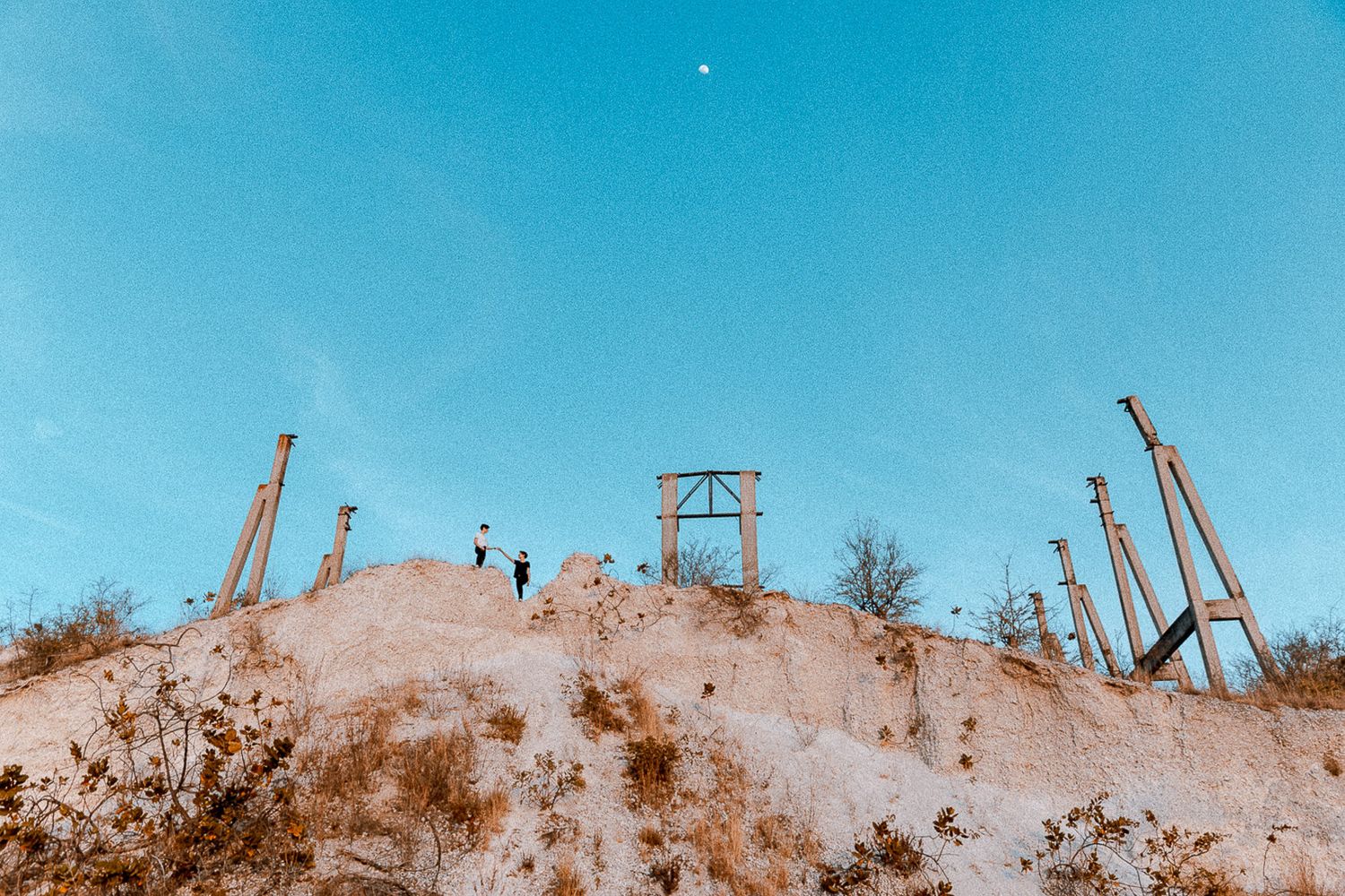 Silhouettes of construction cranes against a bright blue sky atop a sandy hill.