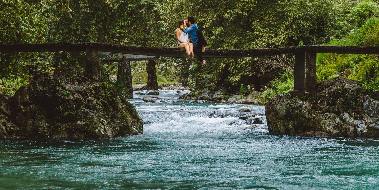 Person sitting on wooden bridge over rushing blue river waters in scenic landscape.