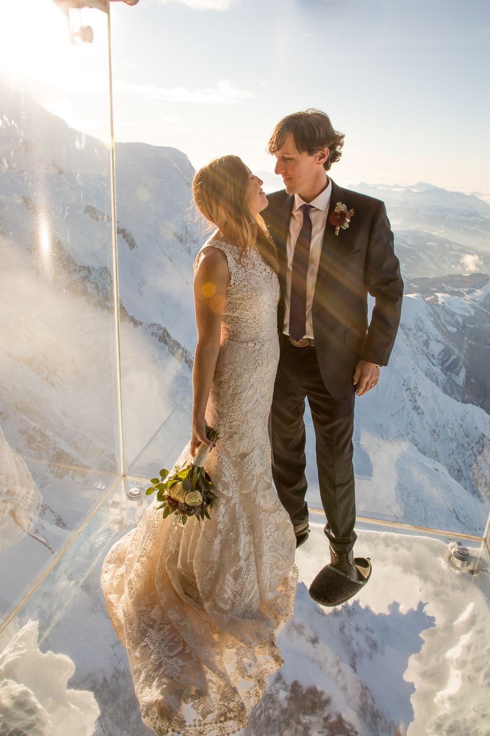 A wedding couple shares an intimate moment atop a snowy mountain peak during a beautiful sunset.