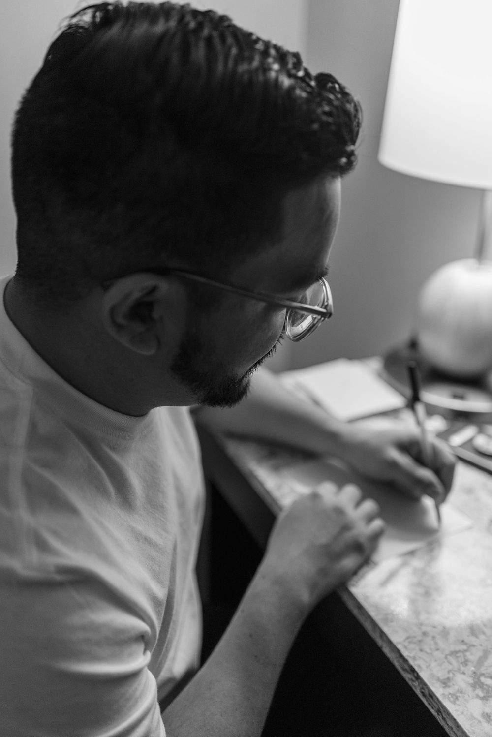 Black and white profile shot of someone writing at a desk with a lamp.