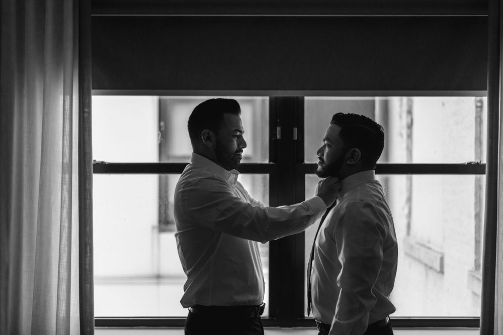 Two men in formal wear stand close together by a window in an intimate black and white moment.