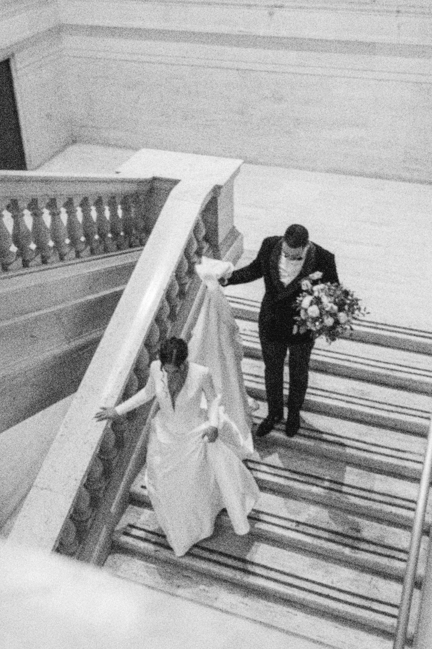 A couple descending an elegant staircase at their wedding, with the bride in white and groom carrying flowers.
