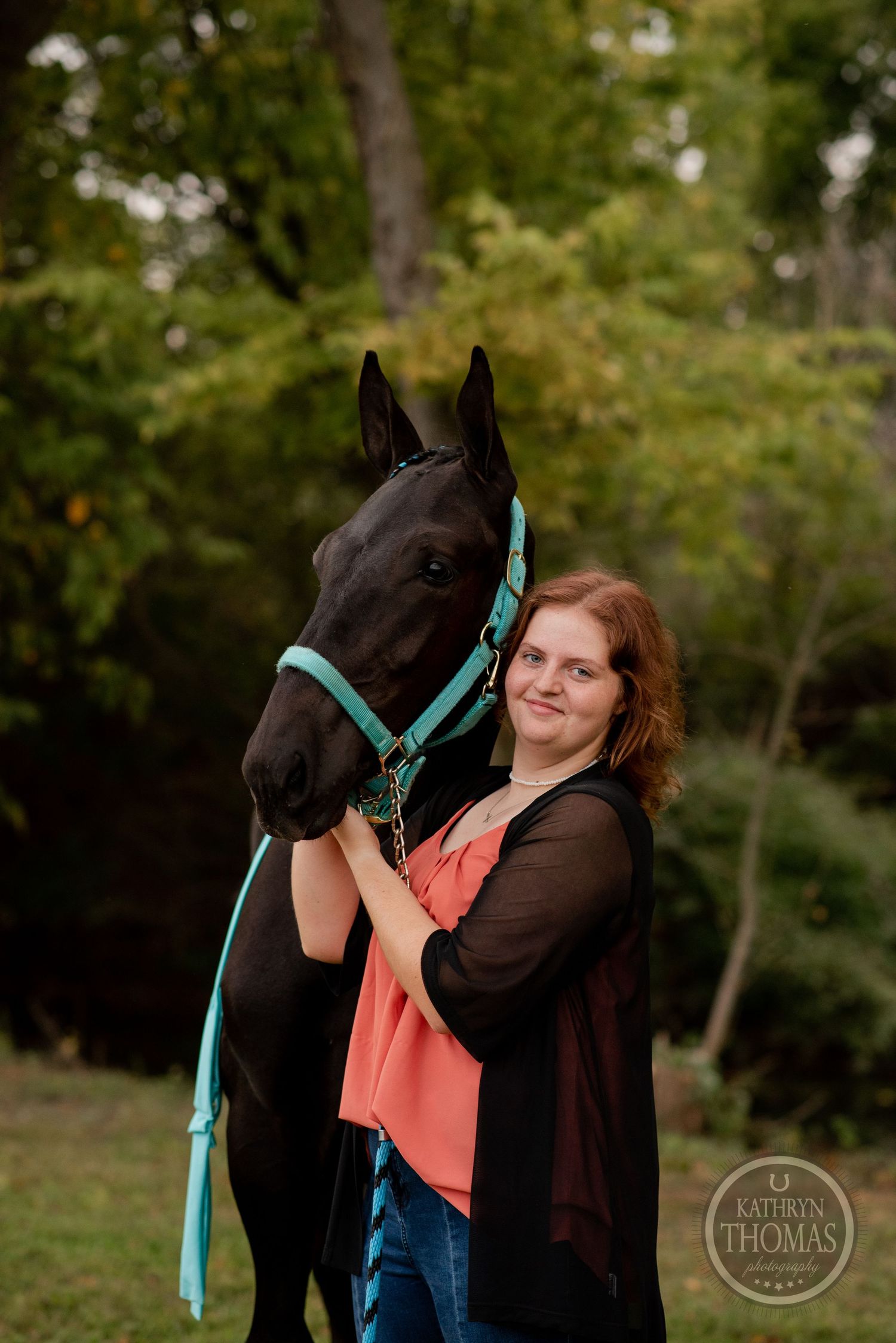 A beautiful black horse wearing a turquoise halter poses outdoors with its handler against a natural wooded background.