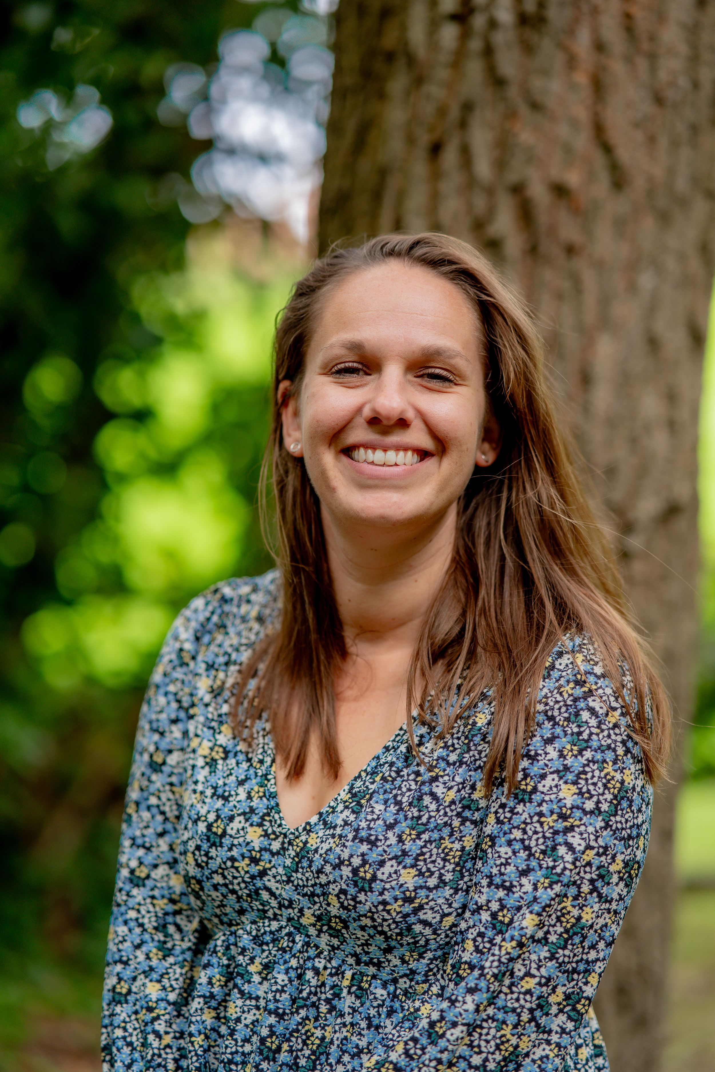 A portrait of someone in a blue floral dress standing against a tree trunk with blurred green foliage in the background.