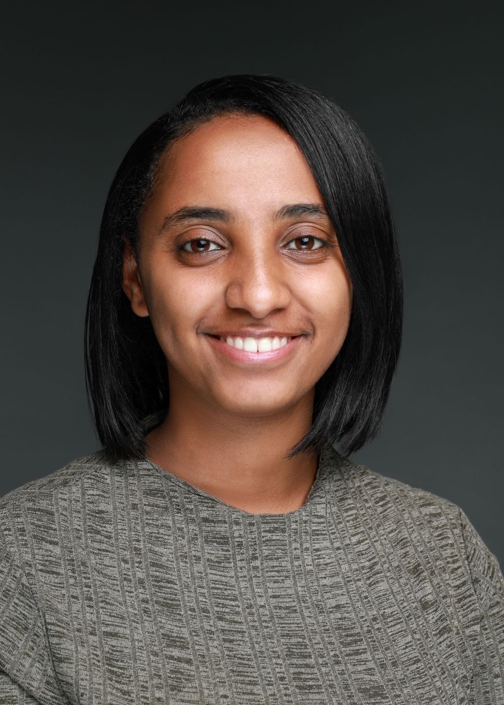 Professional headshot of person with shoulder-length dark hair wearing grey sweater and warm smile.