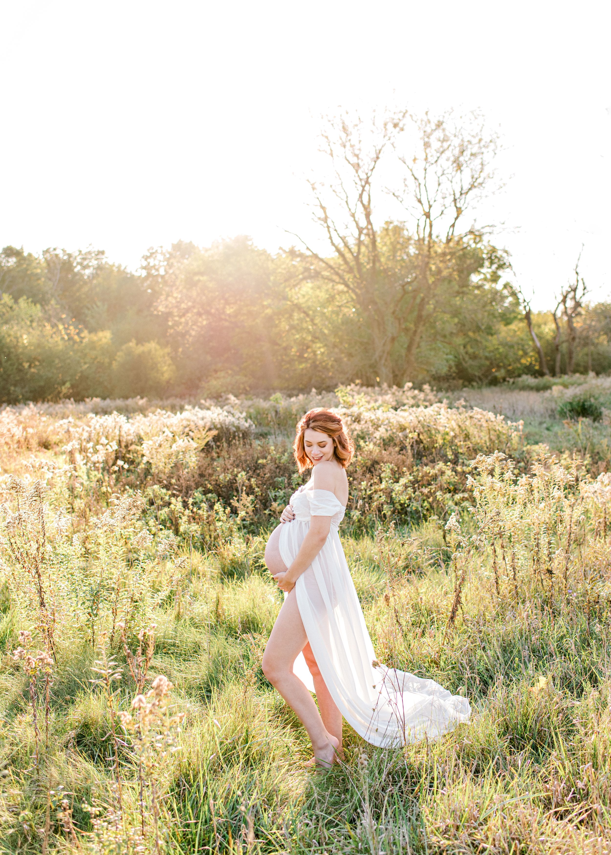 Ethereal maternity photo in flowing white dress during golden hour in field.