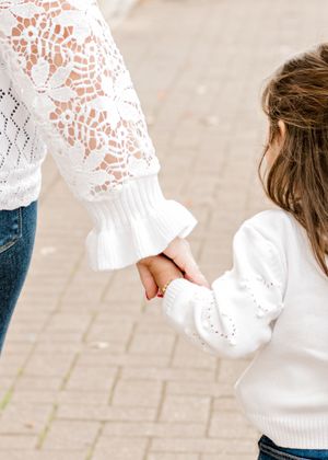 Close-up of holding hands wearing white lace sweaters on stone pathway.