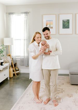 Parents barefoot in bright living room sharing tender moment with newborn by window.