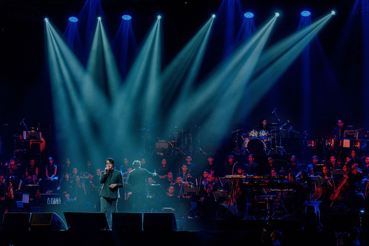 Dramatic blue spotlights illuminate a concert stage with orchestra members performing in the background.