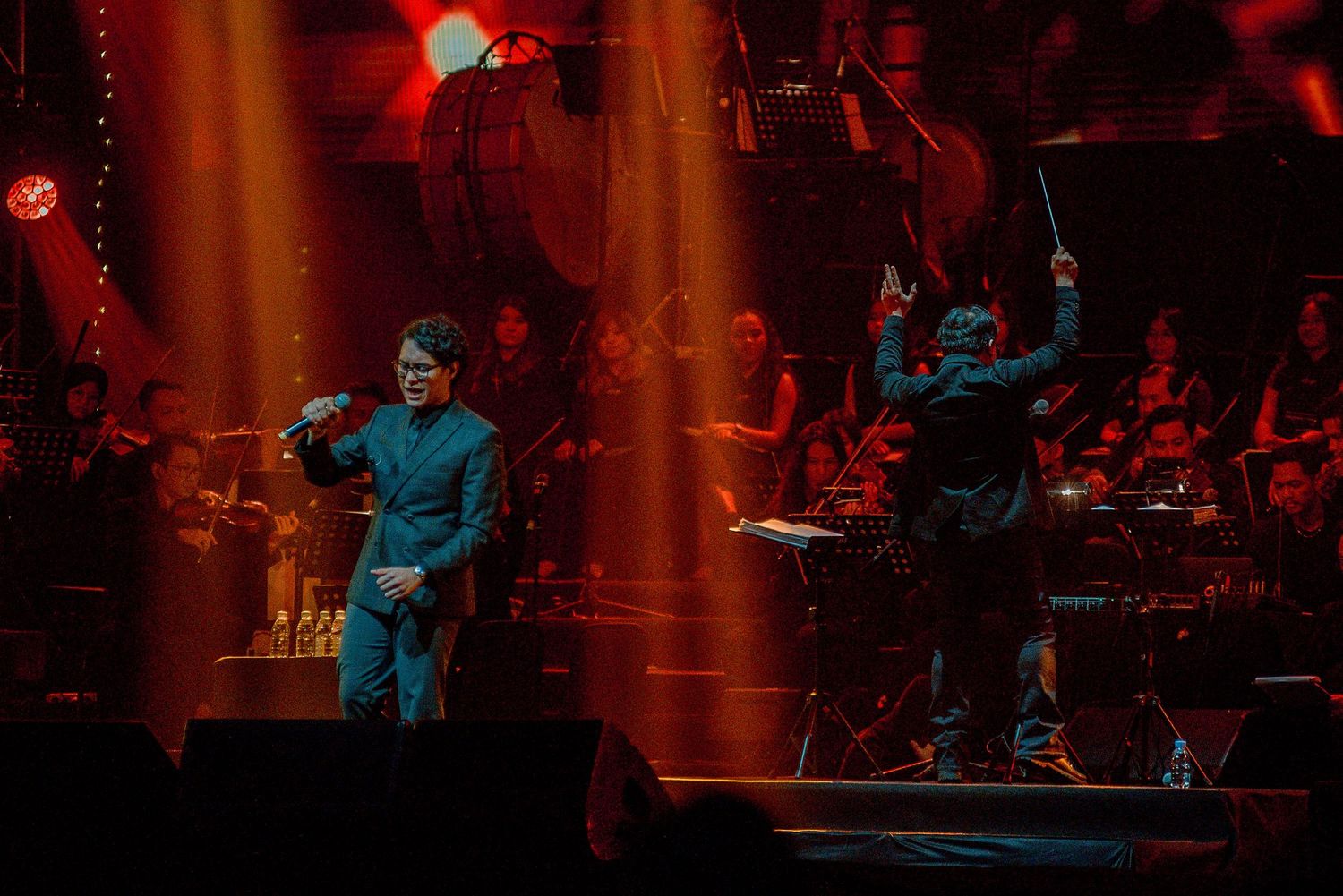 Performers on stage bathed in warm orange lighting with orchestra visible in background.