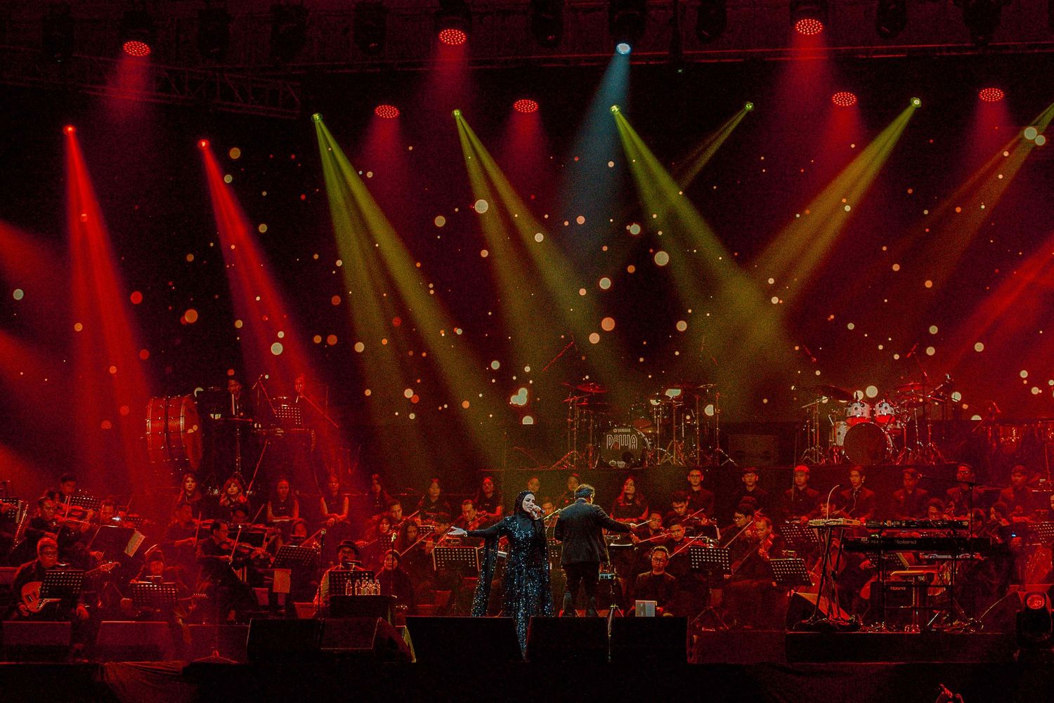Concert stage illuminated by red and yellow spotlights with orchestra performing under dramatic lighting.