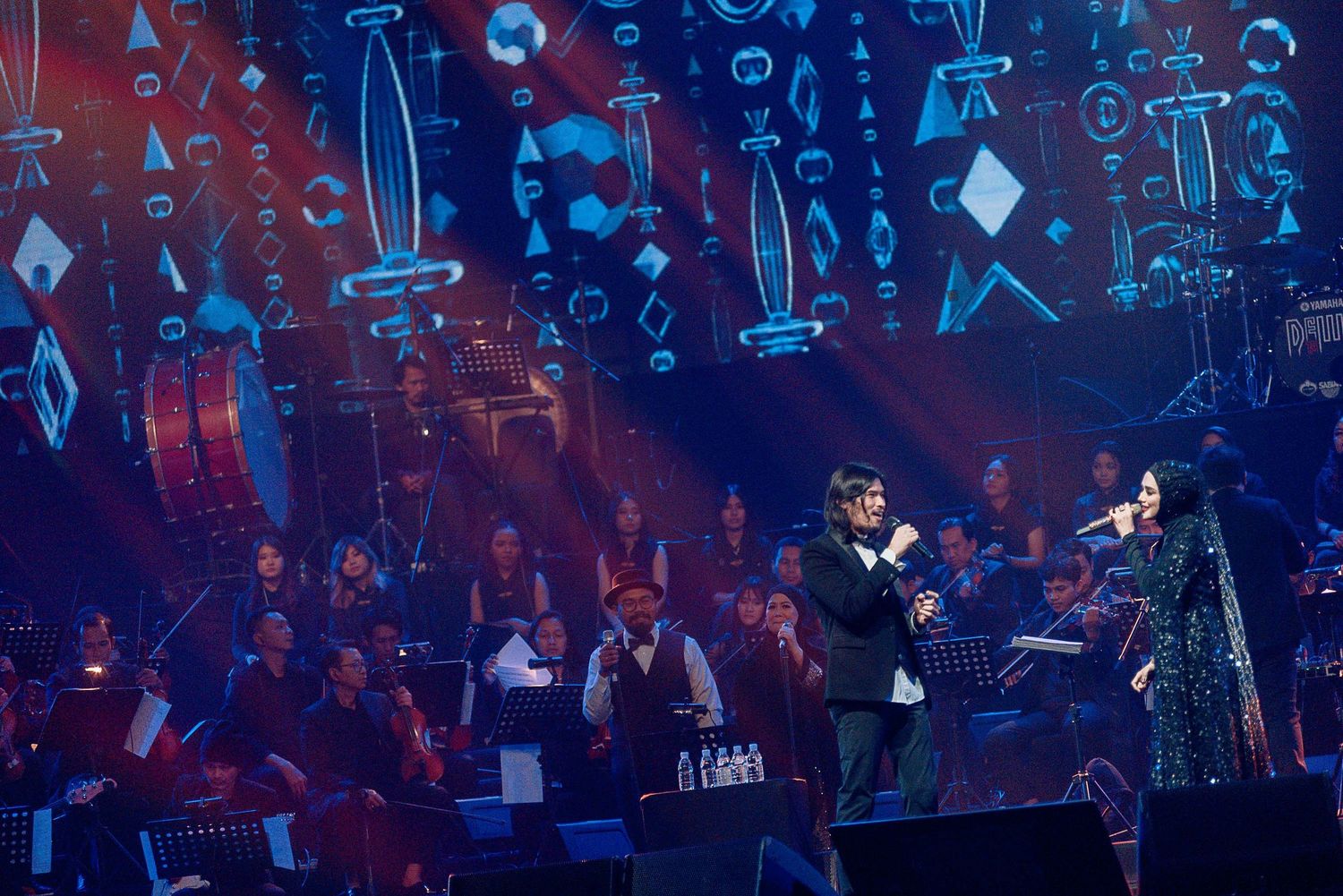 Concert stage with digital geometric projections and performers silhouetted against blue lighting.
