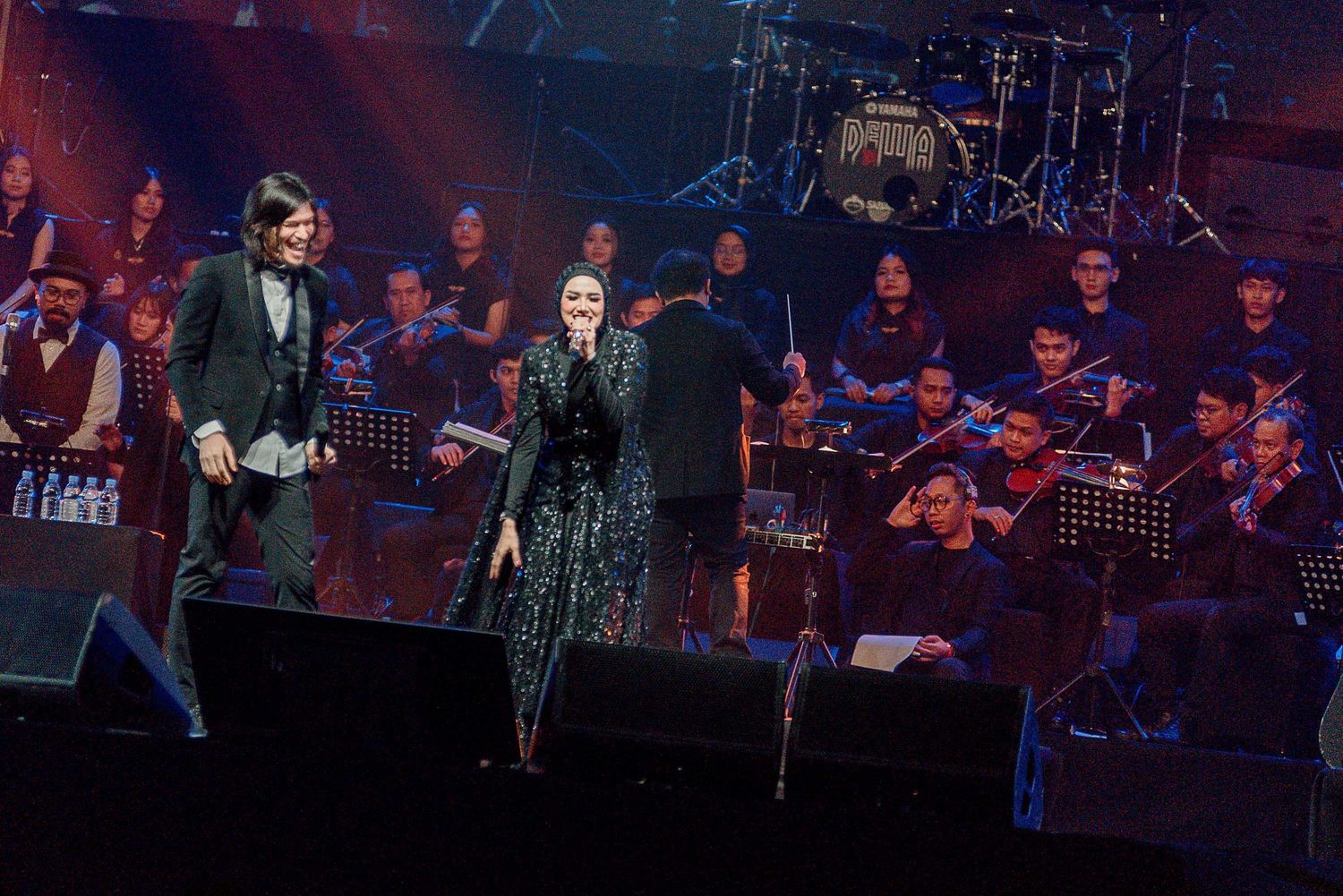 Orchestra performs with featured artists on stage under dramatic concert lighting.