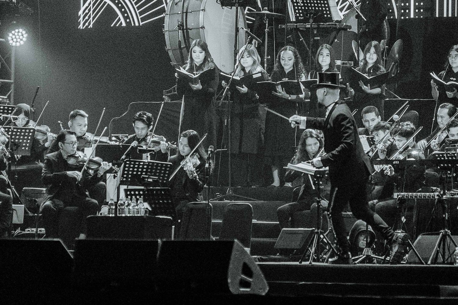 Orchestra performs on stage with dramatic lighting and multiple string instruments in a black and white concert setting.