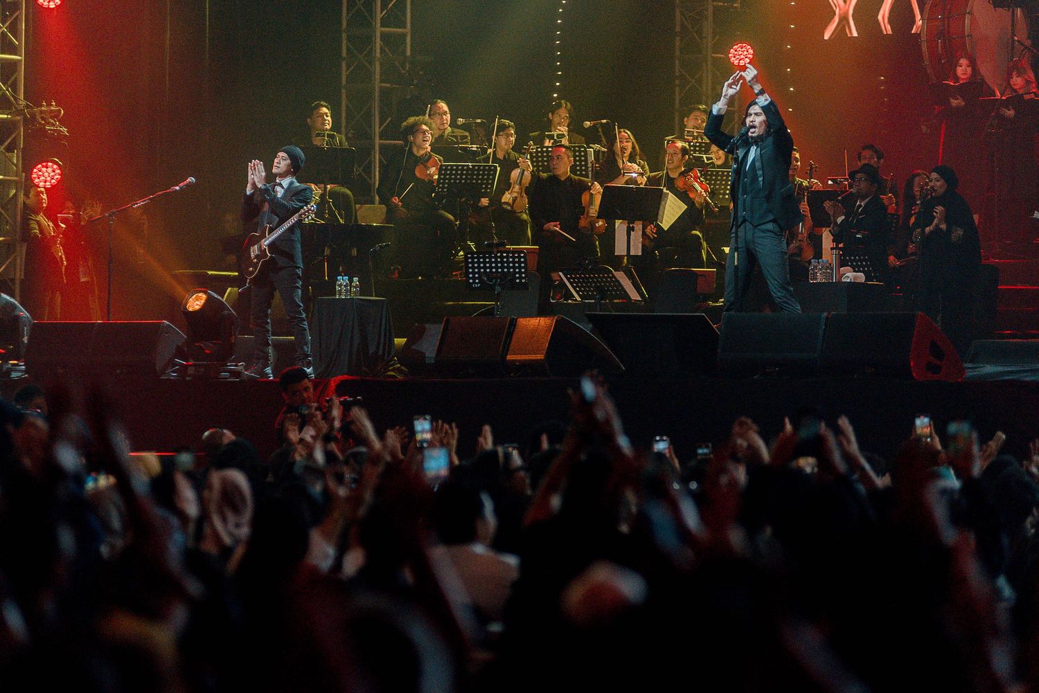 Energetic concert performance with orchestra and crowd raising hands in foreground.