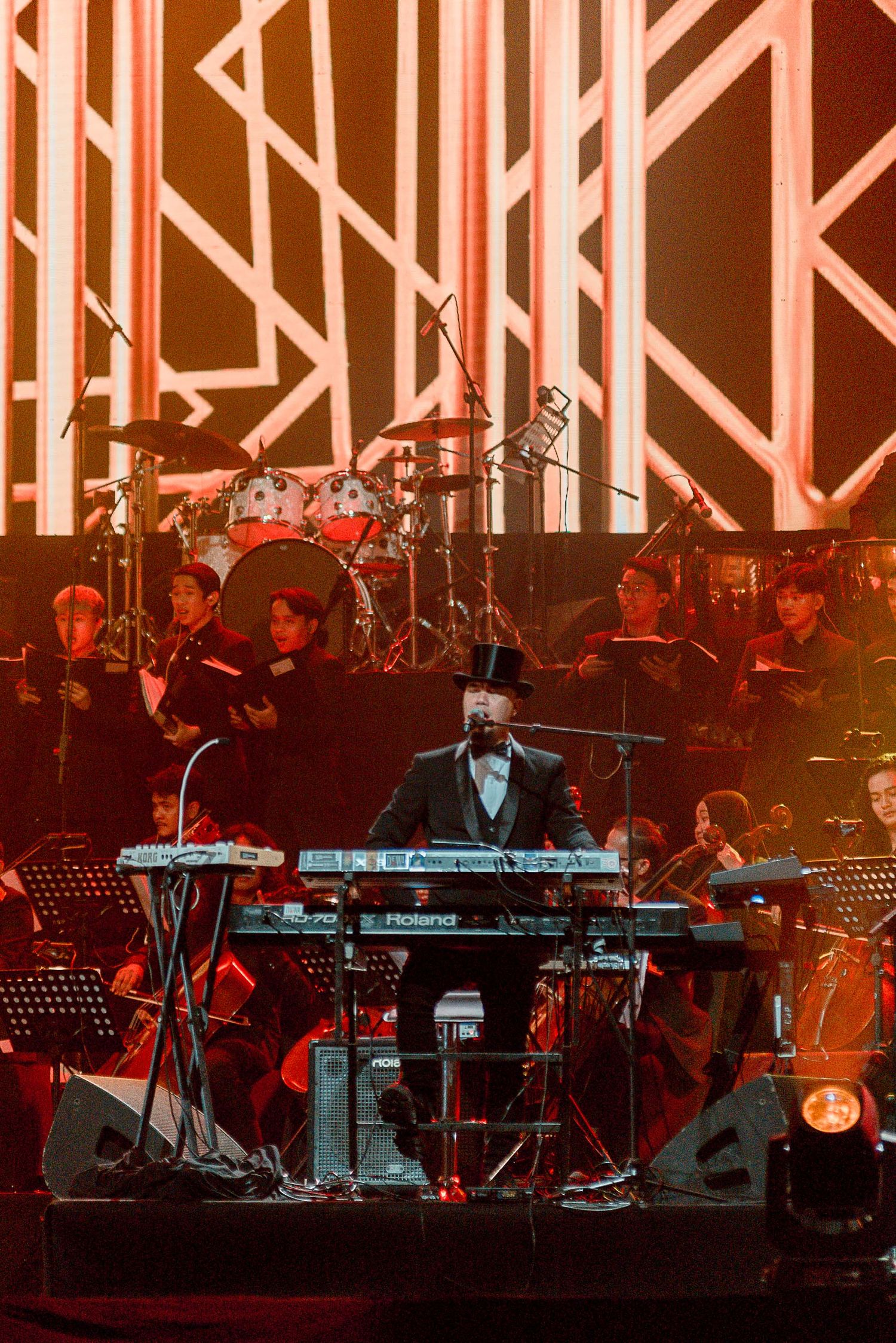 Orchestra performance with geometric orange backdrop and keyboard player in center stage.