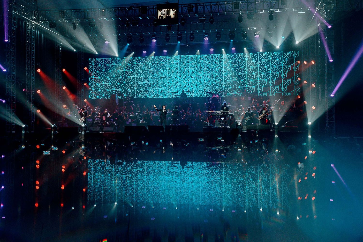 Concert stage with geometric blue light display and reflective floor creating mirror effect.