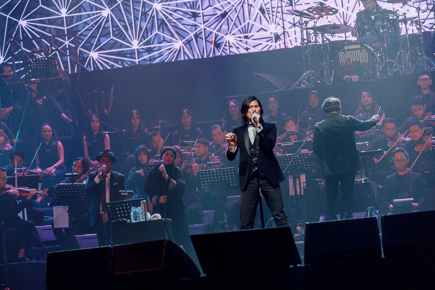 Concert performer on stage with orchestra and large digital backdrop display.