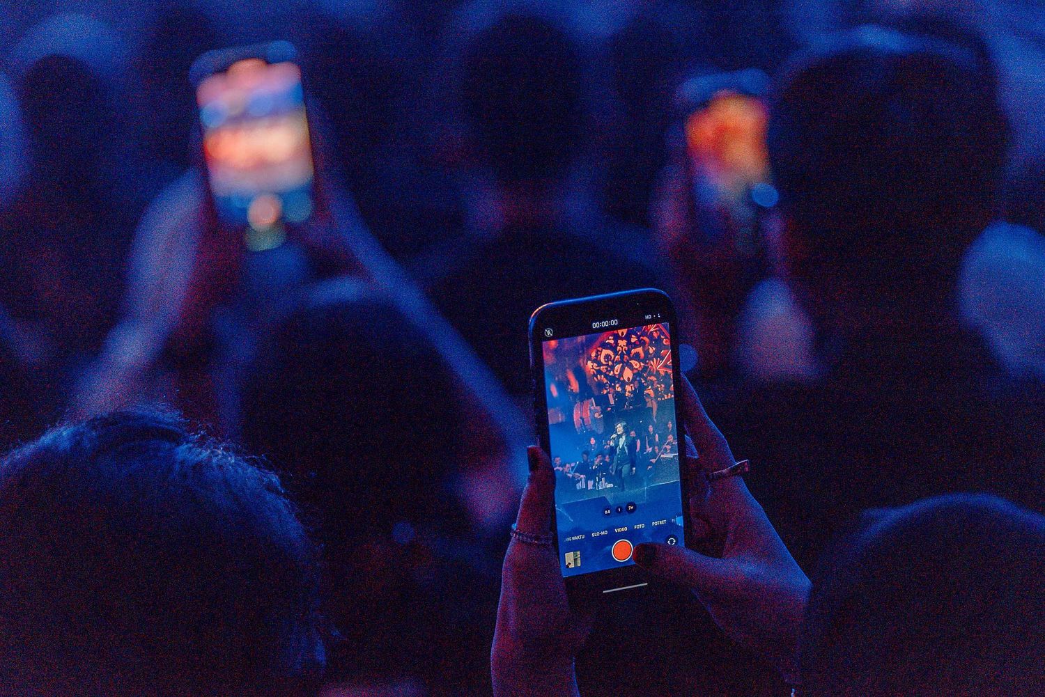 Concert attendee holding up phone to capture performance in blue-tinted crowd.