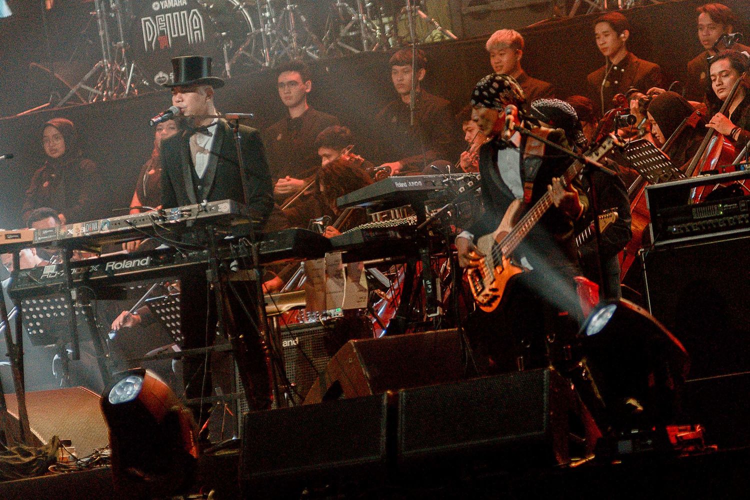Musicians performing with electronic equipment and orchestra on stage with dramatic lighting.