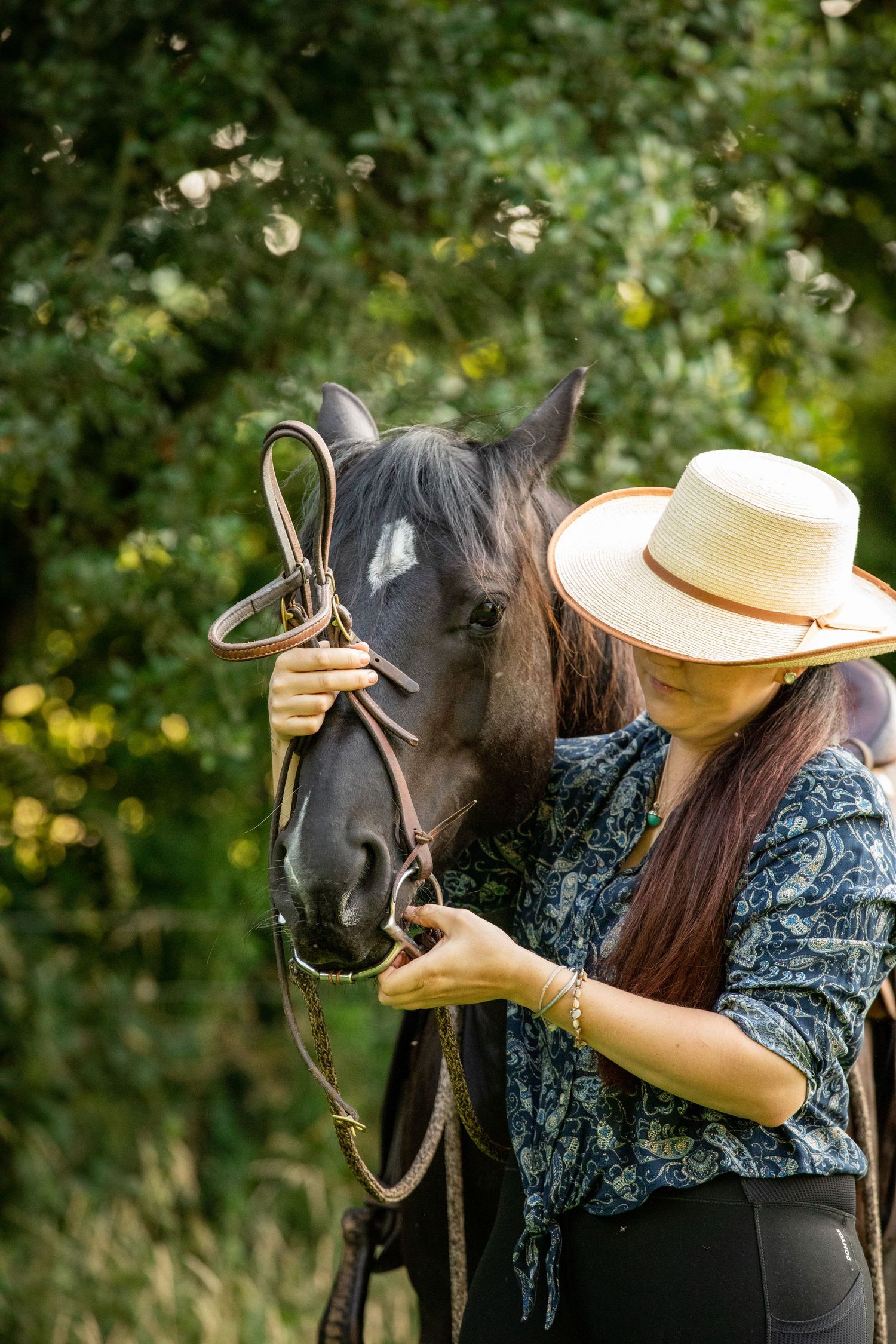 A person in a hat and floral dress adjusts the bridle on a dark horse in dappled sunlight.