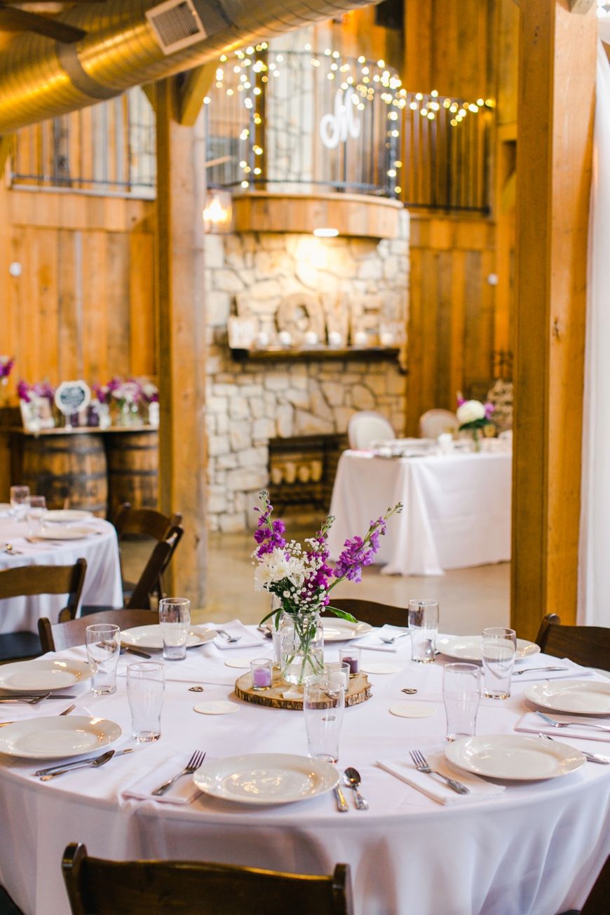 An elegant wedding reception table set with white linens and purple floral centerpiece in a rustic wooden venue.