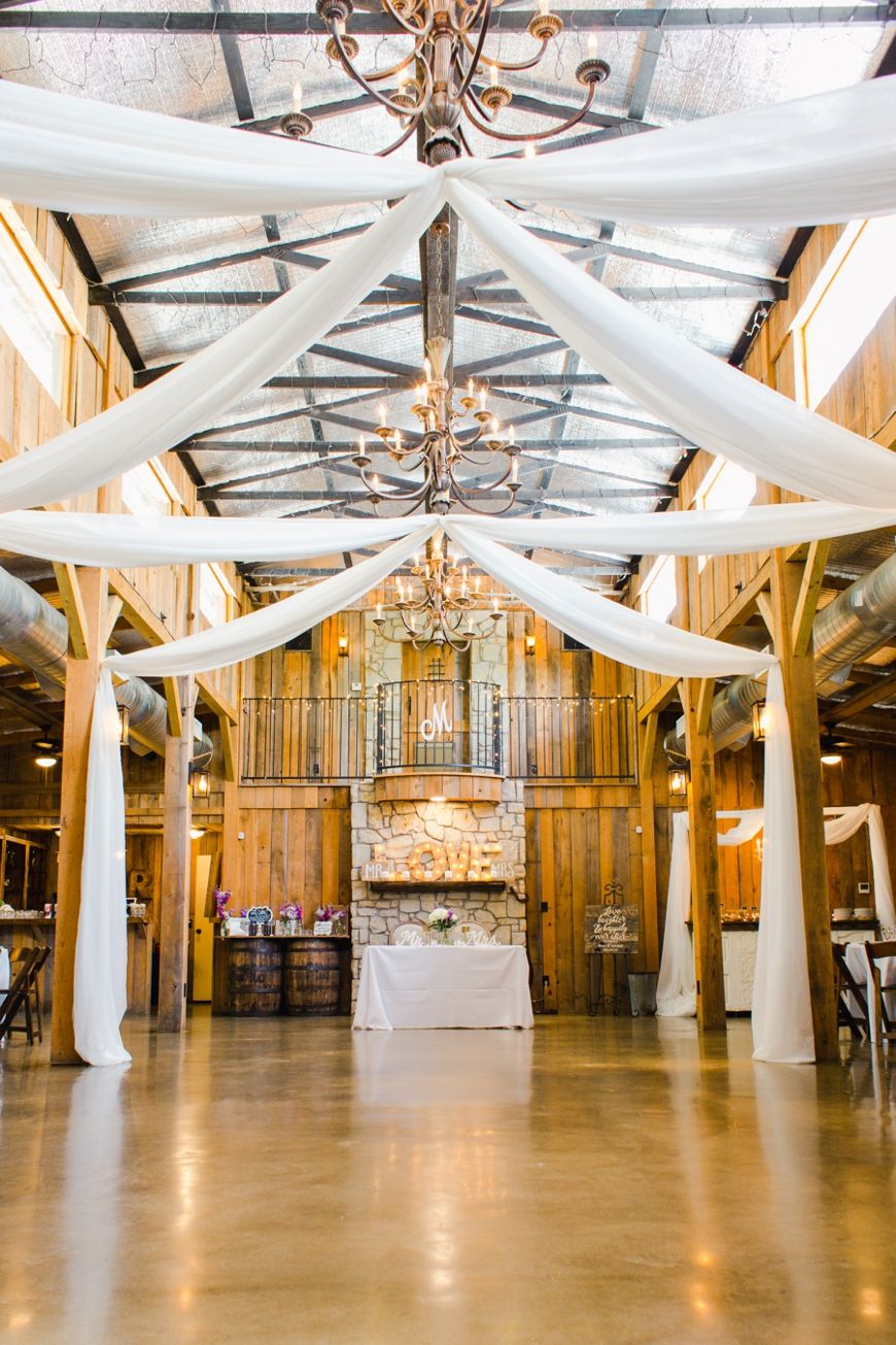 Elegant barn venue interior with white fabric draping and crystal chandeliers hanging from wooden beams.