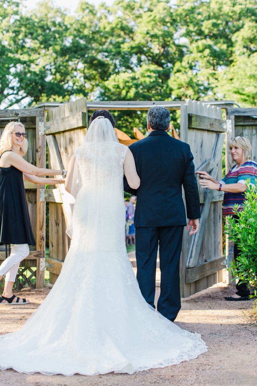 A bride in a flowing white dress walks along a wooden pathway during an outdoor wedding.
