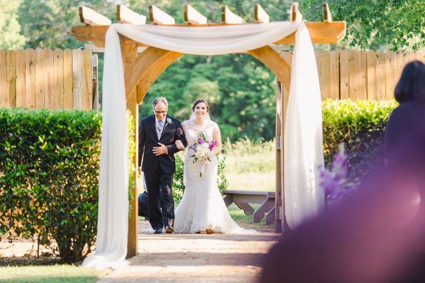 A couple walks through an elegantly draped wooden wedding arch in an outdoor garden ceremony setting.