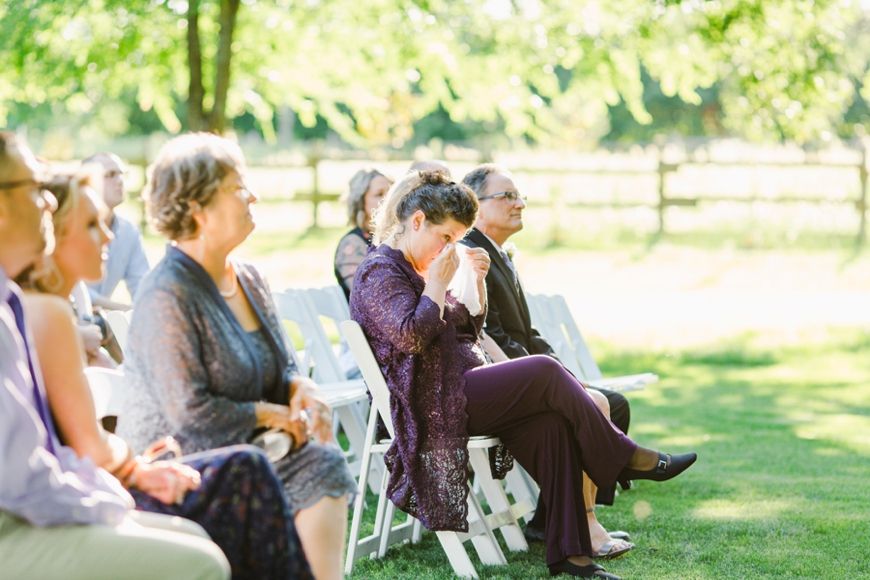 Wedding guests seated on white chairs during an outdoor ceremony in a sunlit garden setting.