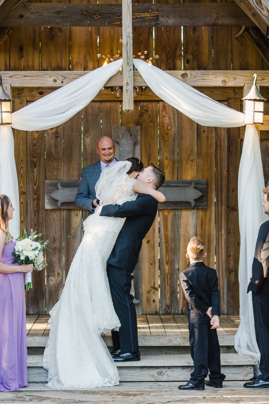A romantic first kiss moment at a rustic outdoor wedding ceremony under draped white fabric.