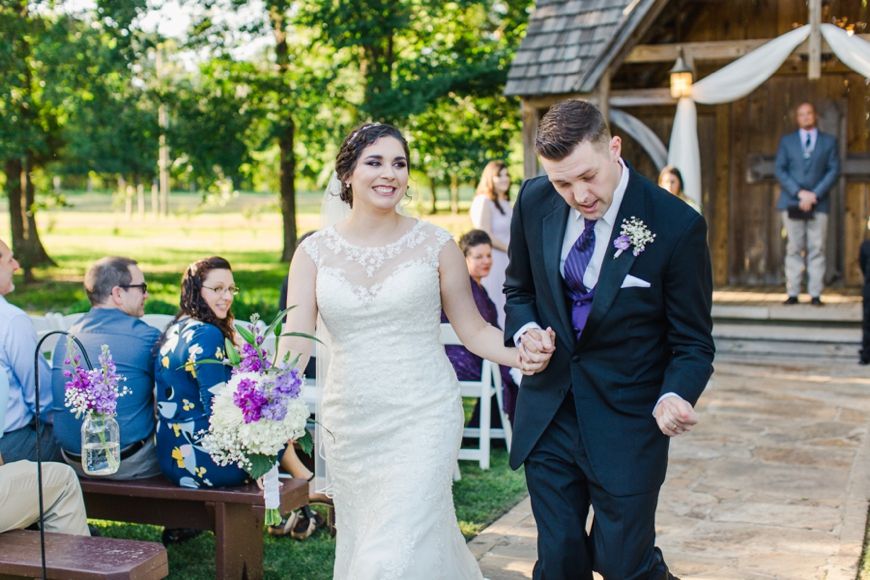 Newlywed couple joyfully walks down aisle after outdoor wedding ceremony with rustic wooden chapel in background.