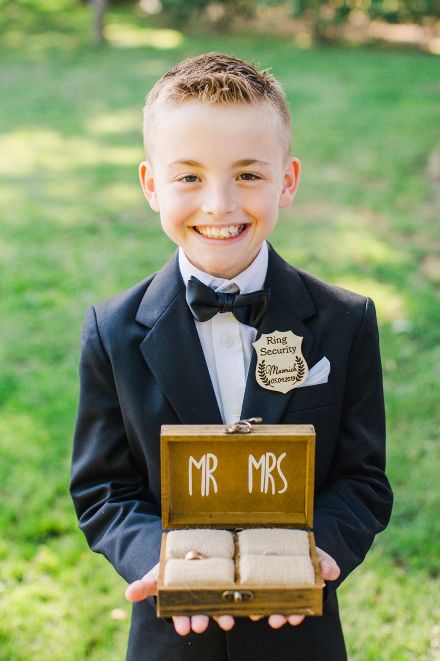 Young ring bearer in tuxedo holds wooden Mr & Mrs box while smiling outdoors.