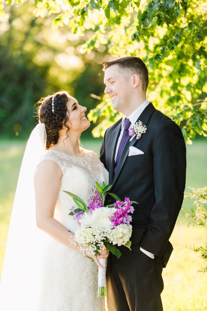 Couple shares intimate moment under tree canopy with bridal bouquet of purple and white flowers.